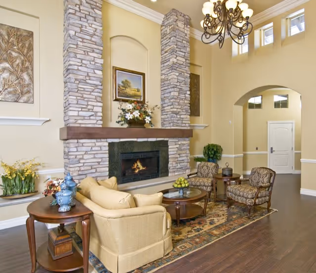 A cozy living room area with a stone fireplace, a beige couch, two patterned armchairs, a wooden coffee table with a bowl of green apples, and a side table with decorative blue vases. The room has high ceilings with a chandelier, light yellow walls, and a patterned rug on dark wooden floors.