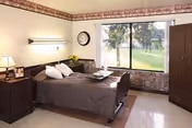 A simple care-center bedroom with a single adjustable bed, nightstand and lamp, clock on the wall, and a large window overlooking a grassy outdoor area.