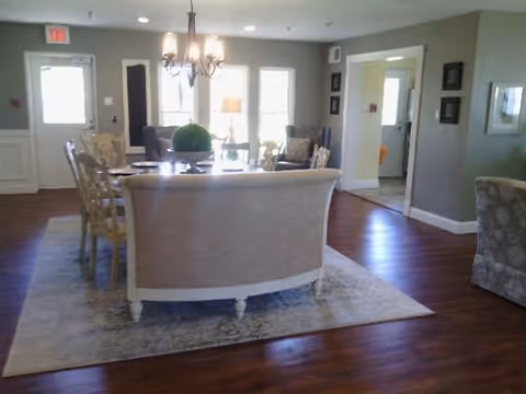 A well-lit living room area with wooden flooring, a light-colored sofa, several chairs around a dining table, a chandelier hanging above the table, and large windows letting in natural light. The walls are painted in a neutral tone and there are framed pictures on the walls.