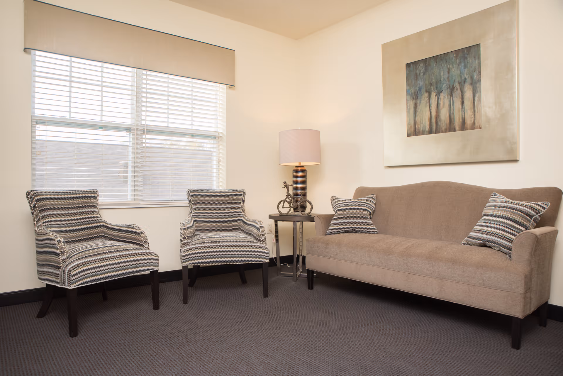 A cozy sitting area with a beige sofa adorned with two striped pillows, two matching striped armchairs, a small round side table with a decorative lamp, and a large window with blinds. A framed painting of trees hangs on the wall above the sofa.