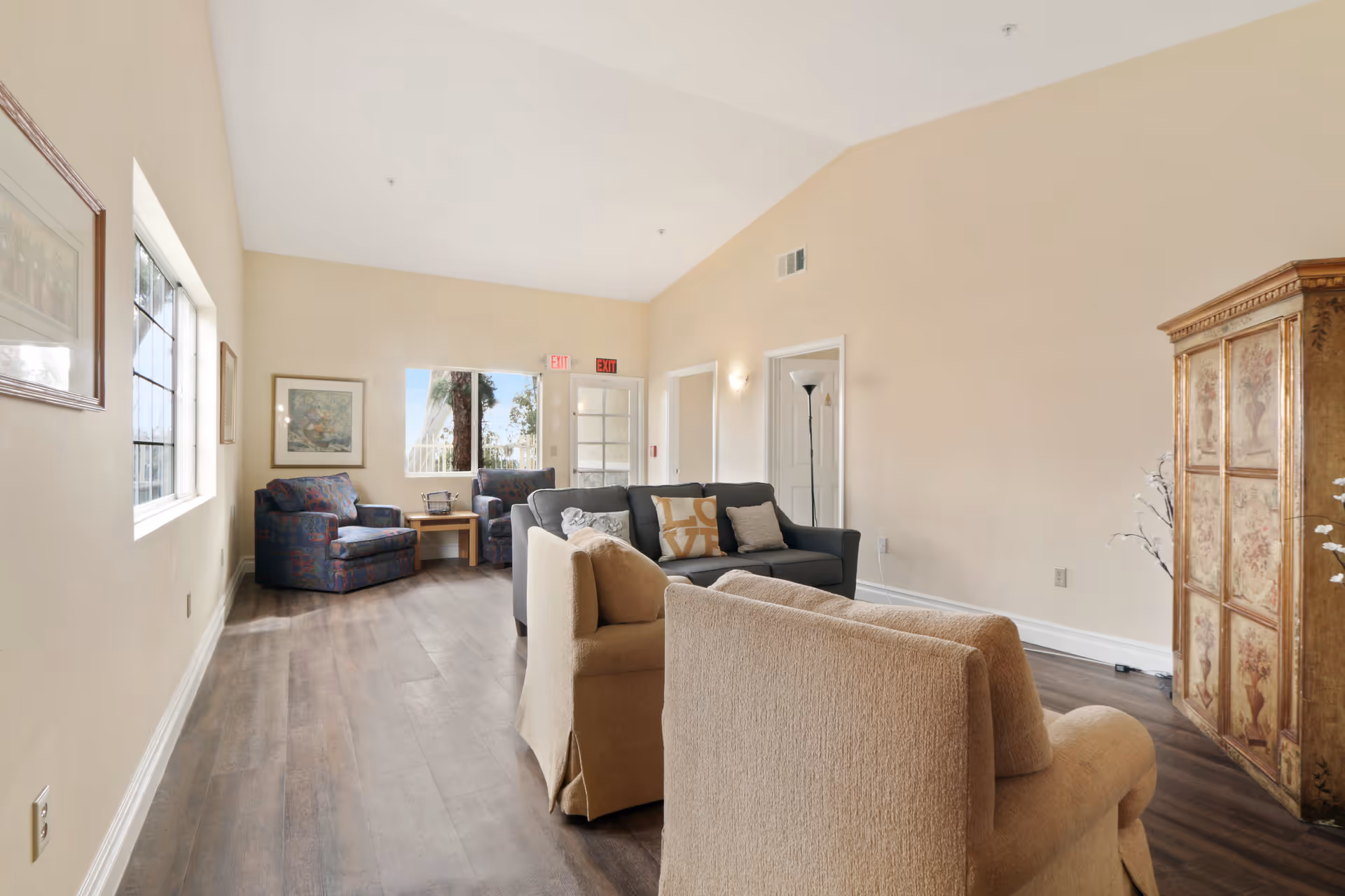 A bright living room with beige walls and wooden flooring featuring a gray sofa with decorative pillows, two beige armchairs, two patterned armchairs near a window, a wooden side table, a tall floor lamp, framed artwork on the walls, and a decorative wooden cabinet with floral designs.