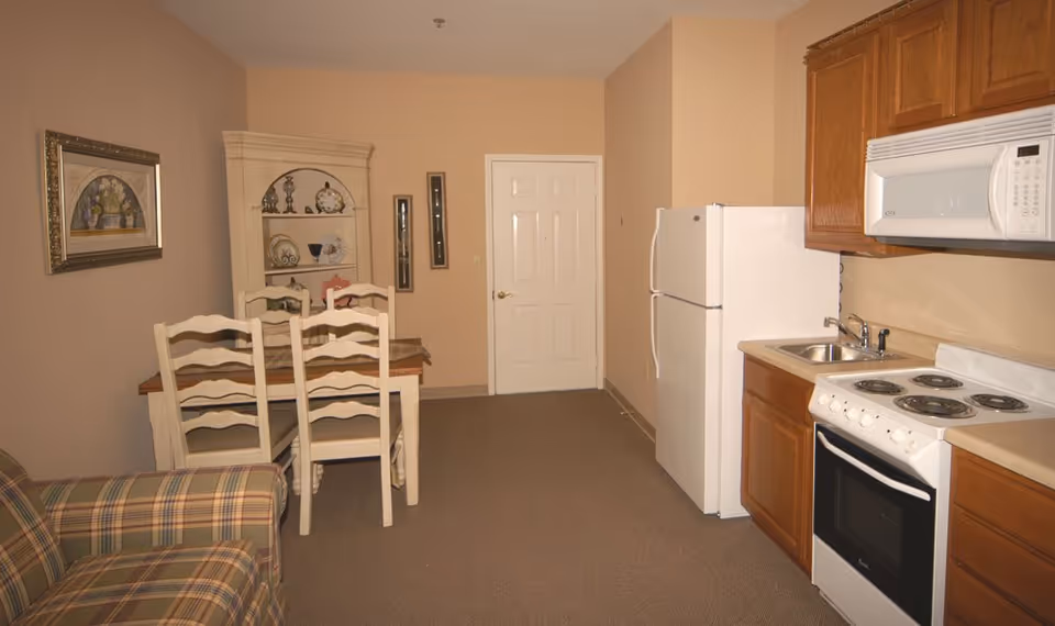 Interior view of a small kitchen and dining area in a senior living facility. The kitchen features wooden cabinets, a white refrigerator, a white microwave, a white electric stove, and a small sink. Adjacent to the kitchen is a dining table with four white wooden chairs. There is a beige wall with a framed painting and a white corner cabinet displaying decorative plates and items. A plaid upholstered armchair is partially visible in the foreground.