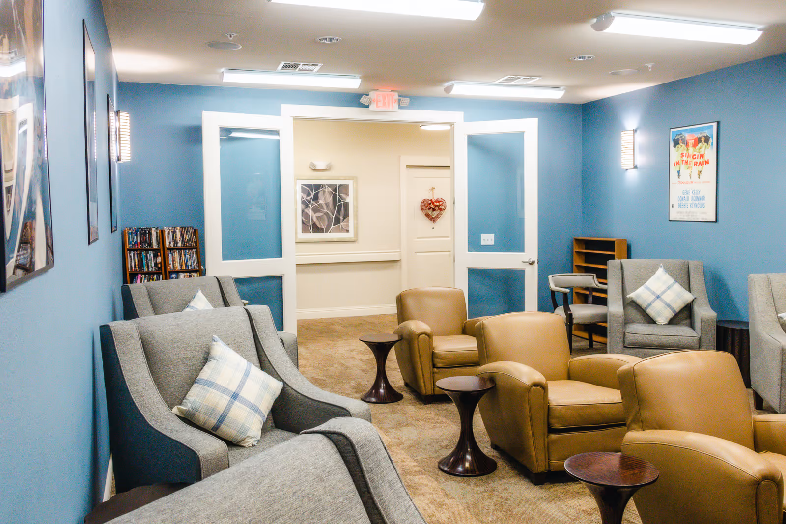 Communal lounge with grey and tan armchairs, small side tables, blue walls, framed artwork and a doorway.