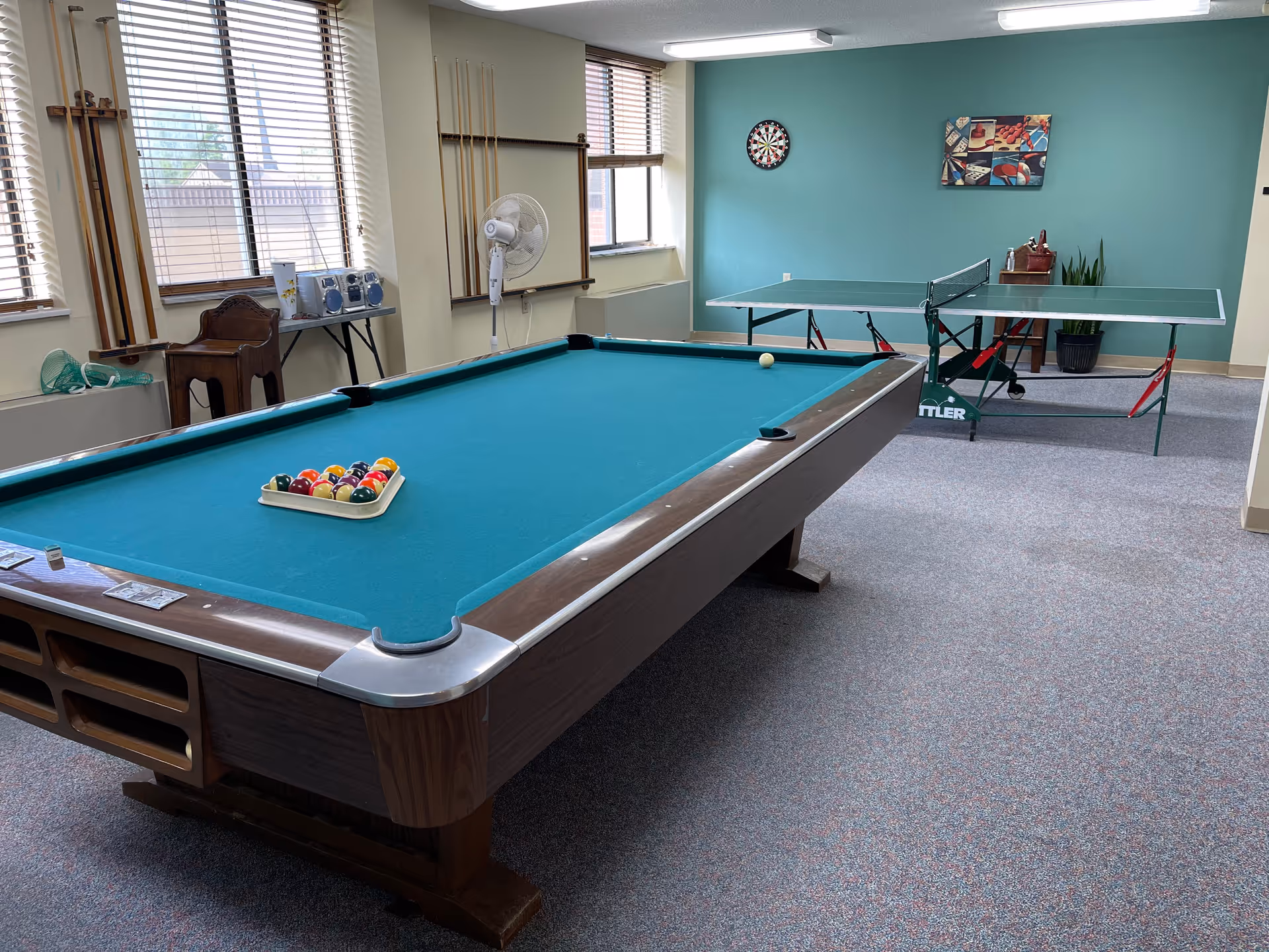A recreational room with a pool table in the foreground, set up with billiard balls and a cue ball. In the background, there is a ping pong table, a dartboard mounted on a teal-colored wall, and a colorful painting. The room has windows with blinds, a standing fan, a small wooden chair, and a table with a stereo system and other items.
