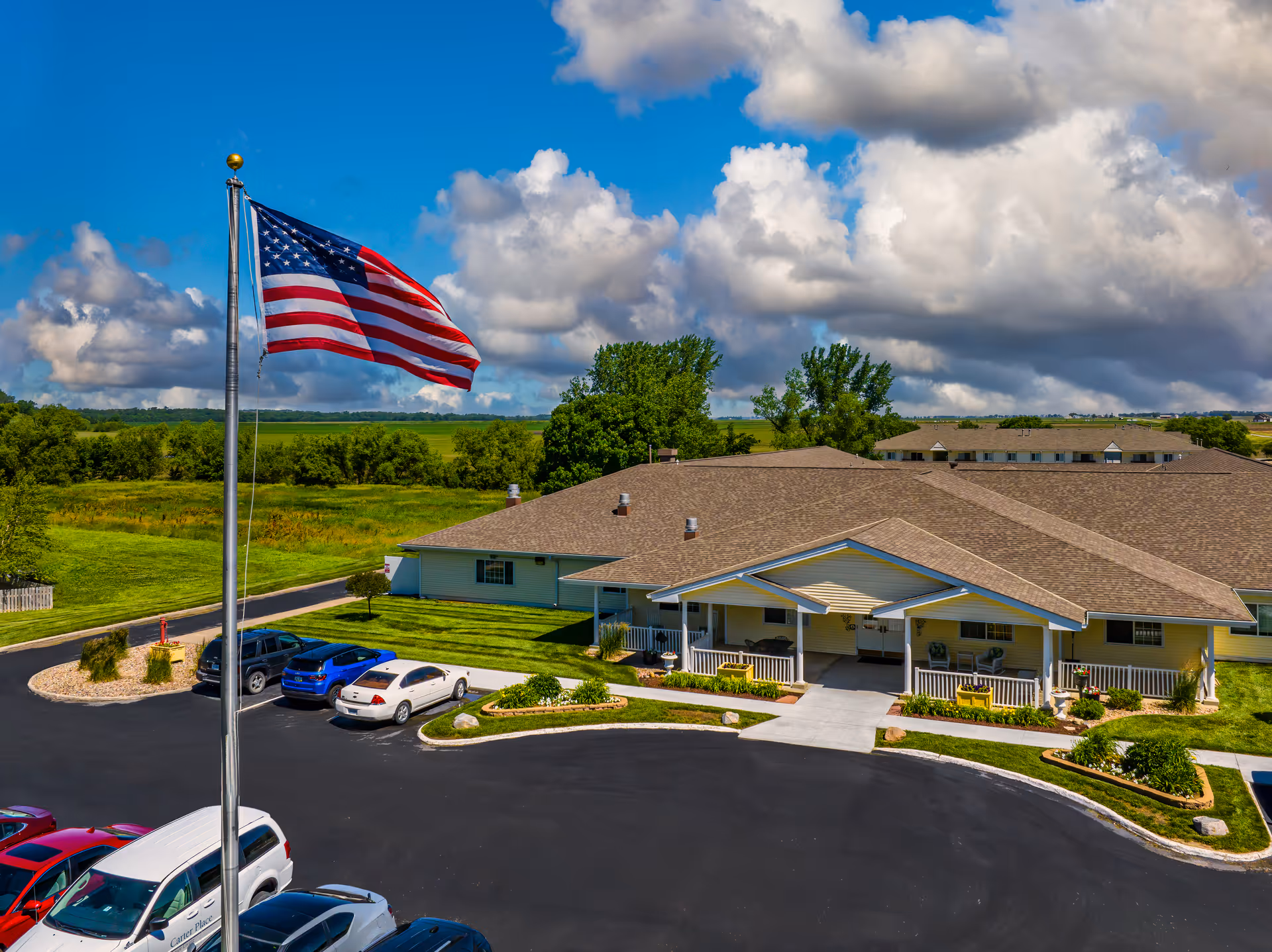 Aerial view of Carter Place Senior Living facility showing a single-story building with a covered entrance, surrounded by a parking lot with several cars parked. An American flag on a tall flagpole is prominently visible in the foreground. The background features green fields, trees, and a partly cloudy blue sky.
