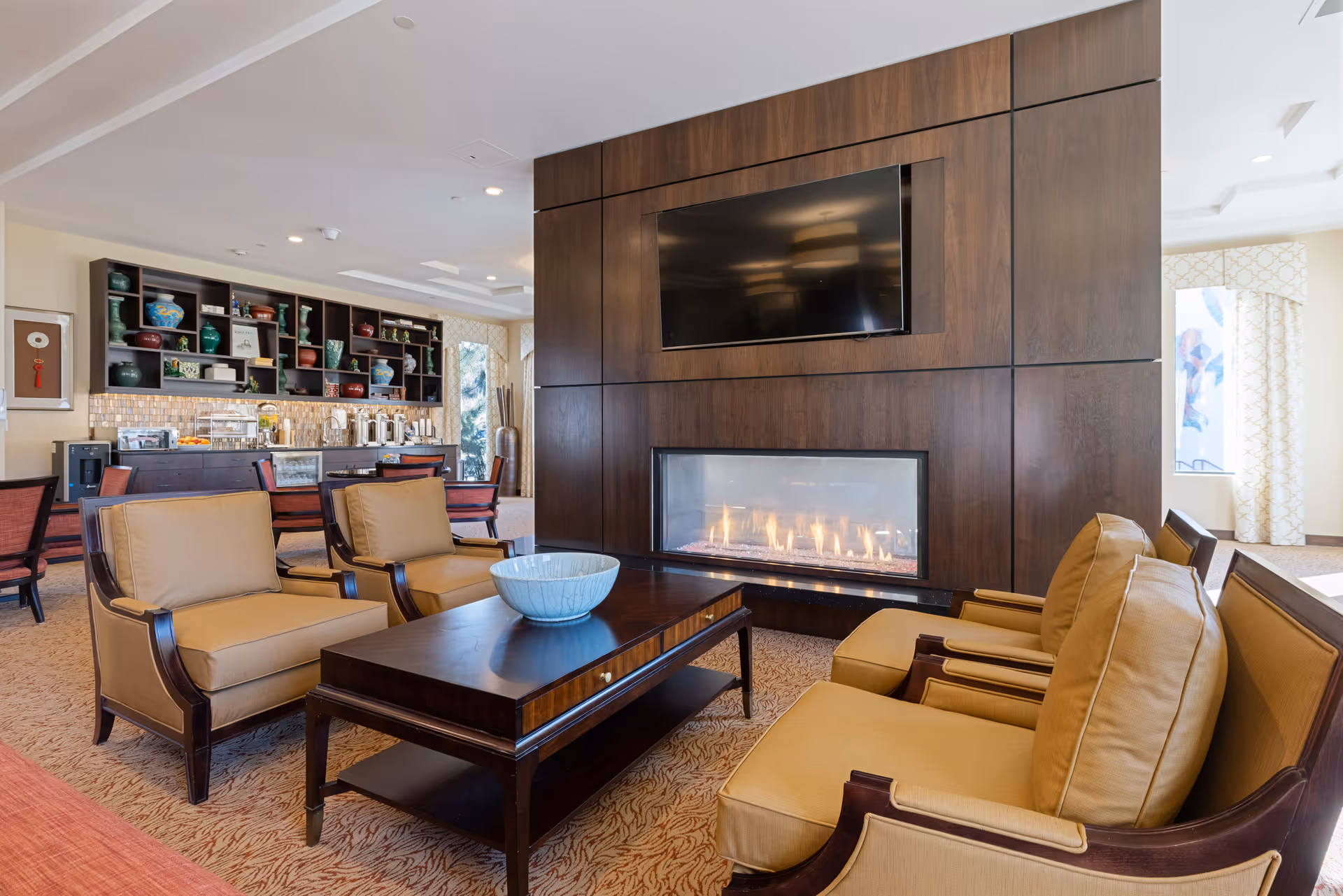 Cozy lounge with armchairs and a coffee table facing a wall-mounted TV above a linear fireplace, with a kitchenette and shelving in the background.