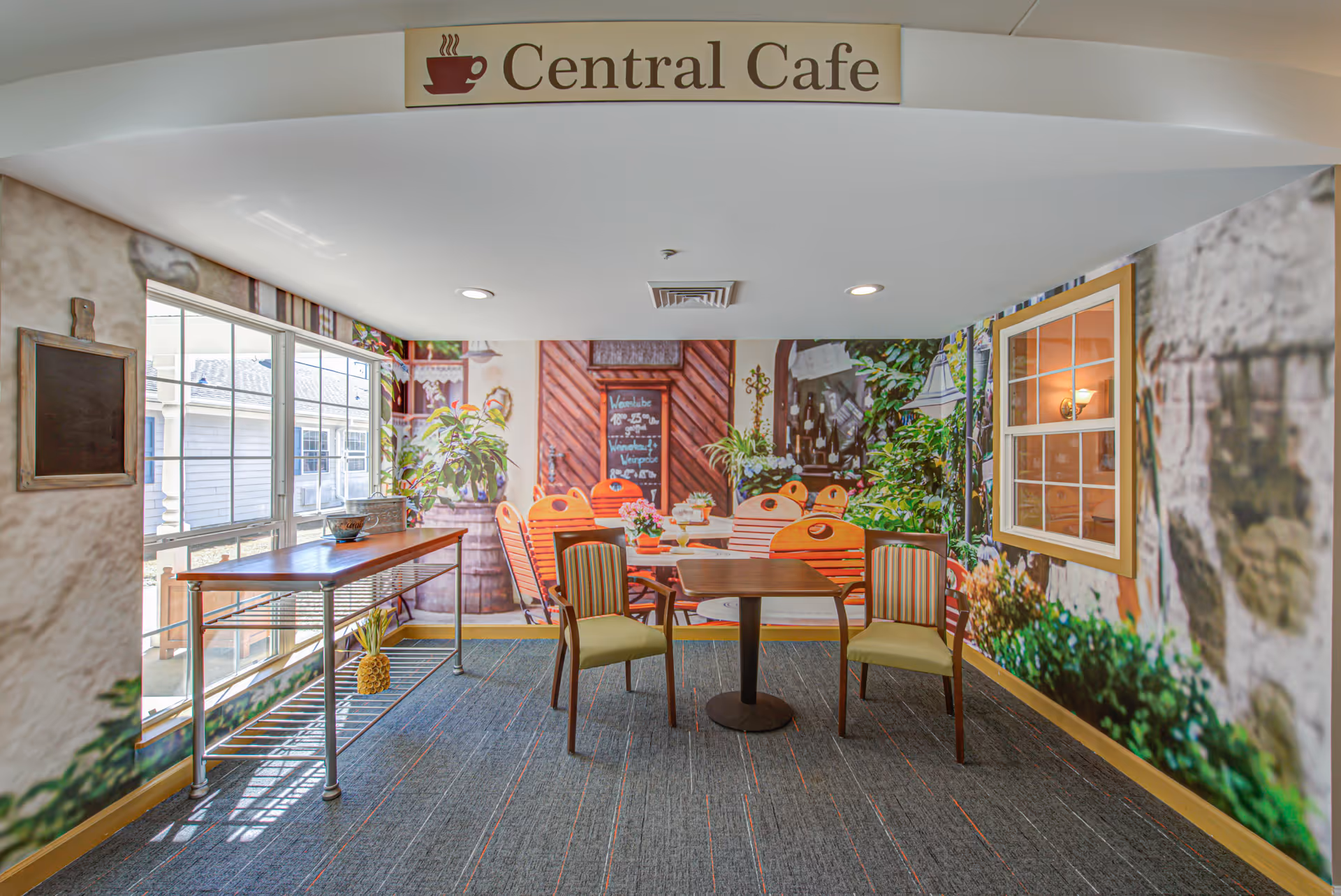 Indoor seating area labeled Central Cafe with two chairs and a small table in front of a wall mural depicting an outdoor cafe scene with plants and chairs. A long table is positioned near a window on the left side.