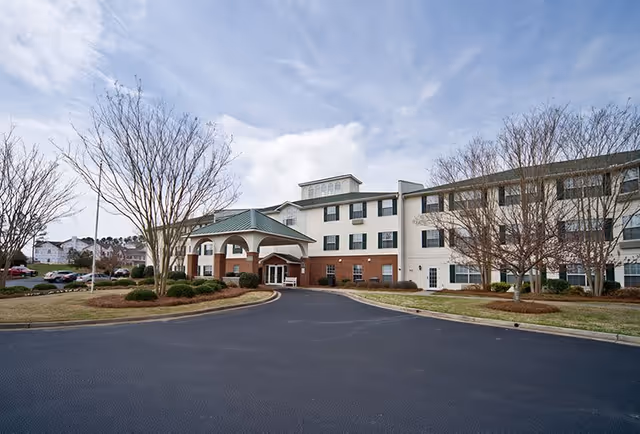 Exterior view of a three-story senior living facility building with a covered entrance, surrounded by leafless trees and a paved driveway under a partly cloudy sky.