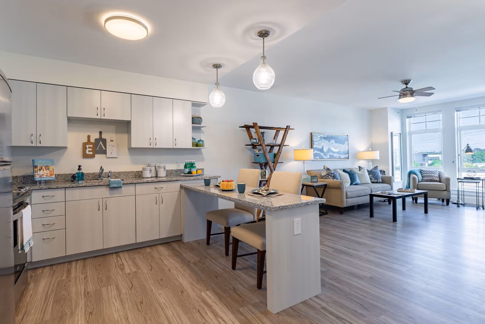 Bright and modern open-concept living space featuring a kitchen with light wood cabinets, granite countertops, and two pendant lights above a breakfast bar with two chairs. The living room area has a beige sofa with multiple blue and white pillows, a matching armchair, a coffee table, a wooden shelf, two table lamps, and large windows letting in natural light.