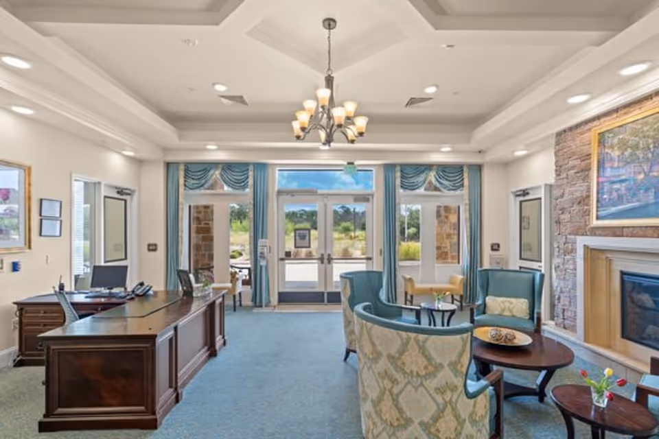 A bright and spacious reception area in a senior living facility with a wooden desk and computer on the left, comfortable upholstered chairs and sofas arranged around coffee tables on the right, a stone fireplace with a framed painting above it, large windows with blue curtains letting in natural light, and a chandelier hanging from the ceiling.