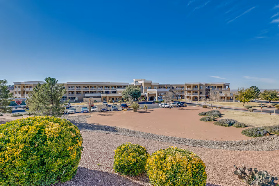 Wide exterior view of Solstice Senior Living at Las Cruces, showing a large three-story building with balconies, a parking lot with several cars, landscaped bushes, and a clear blue sky.