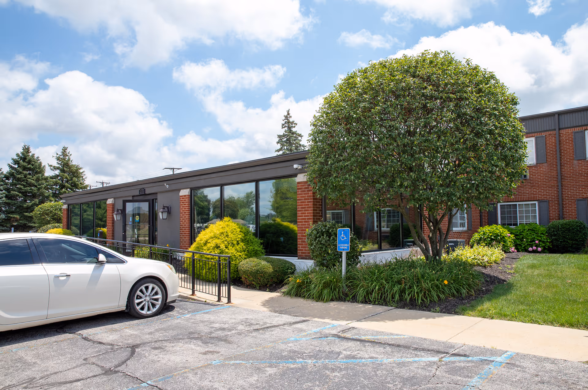 Exterior view of a single-story brick and glass building with a wheelchair accessible ramp, surrounded by green shrubs and a tree, with a white car parked in front under a partly cloudy sky.