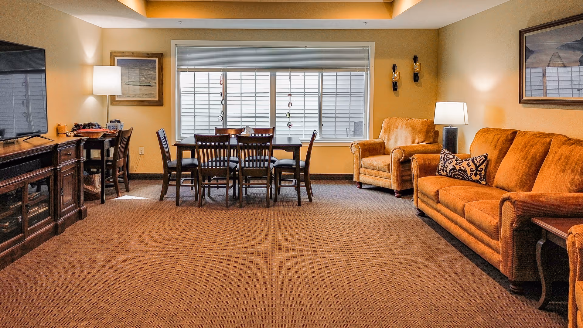 A spacious communal living room with orange sofas and armchairs, a dining table by a large window, and a television console.