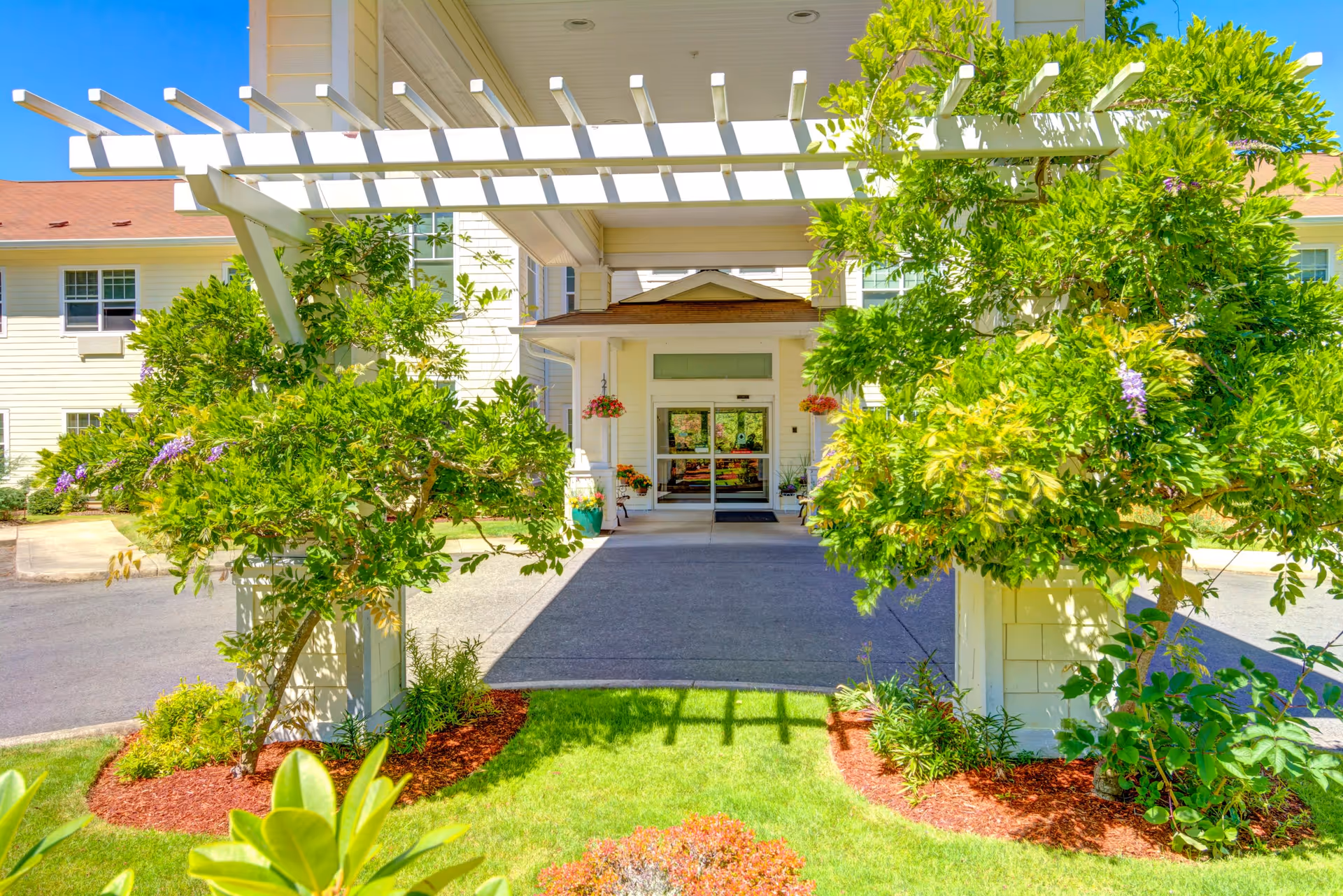 Entrance to a senior living facility with a white pergola covered in green vines and plants, a paved driveway, and a building with glass doors in the background under a clear blue sky.