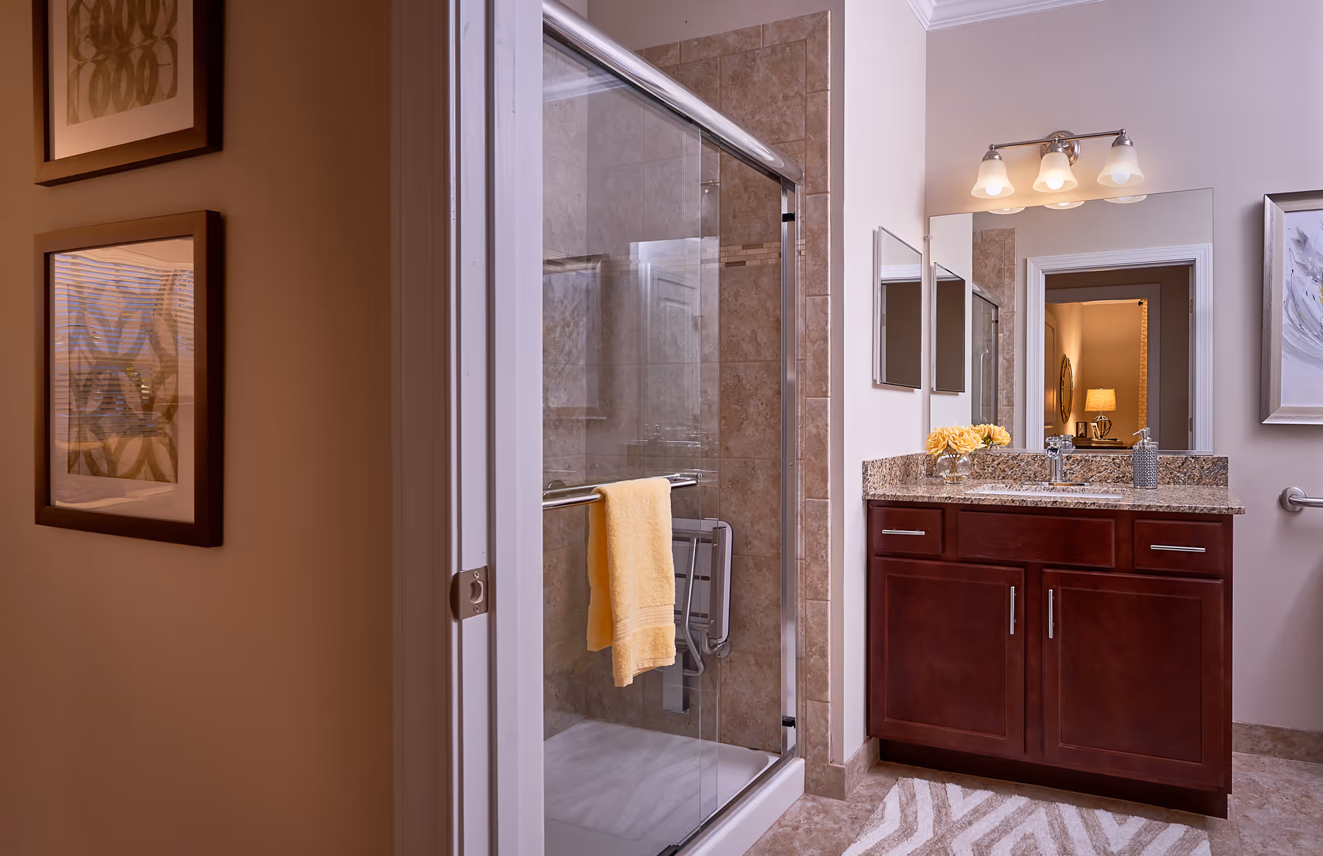A modern bathroom with a glass-enclosed shower featuring beige tiles and a yellow towel hanging on the shower door. There is a dark wood vanity with a granite countertop, a sink, a mirror, and three light fixtures above it. The bathroom has neutral-colored walls and a patterned rug on the floor. Two framed artworks are visible on the adjacent wall outside the bathroom.