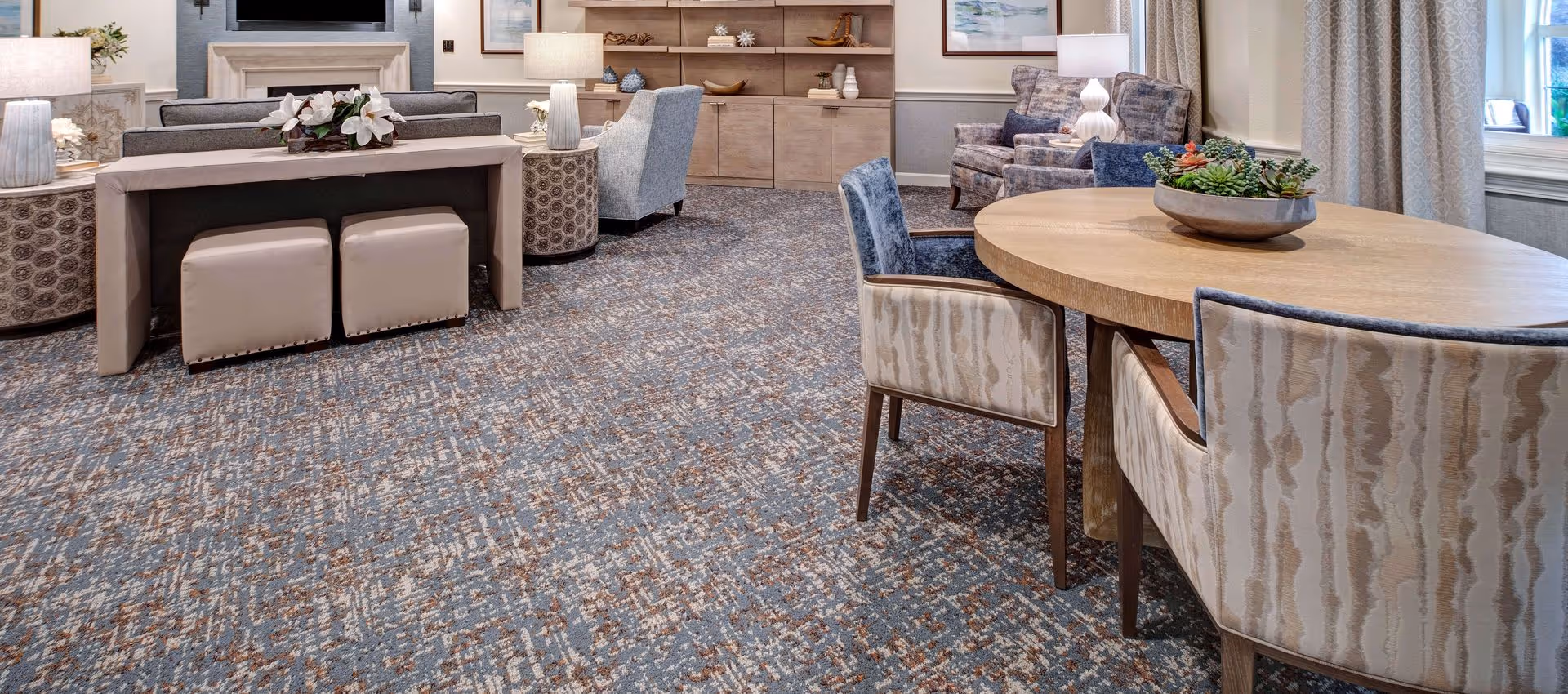 Communal lounge with a round wooden table and upholstered chairs, sofas, ottomans and a TV in a senior living facility.