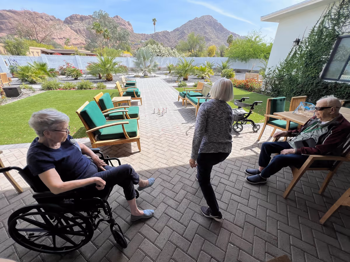 Three elderly individuals in an outdoor patio area of a senior living facility. One person is seated in a wheelchair on the left, another person is standing in the center, and a third person is seated on a chair on the right. The patio has green cushioned wooden chairs arranged along a paved walkway leading to a garden with various plants and palm trees. Mountains and a clear blue sky are visible in the background.