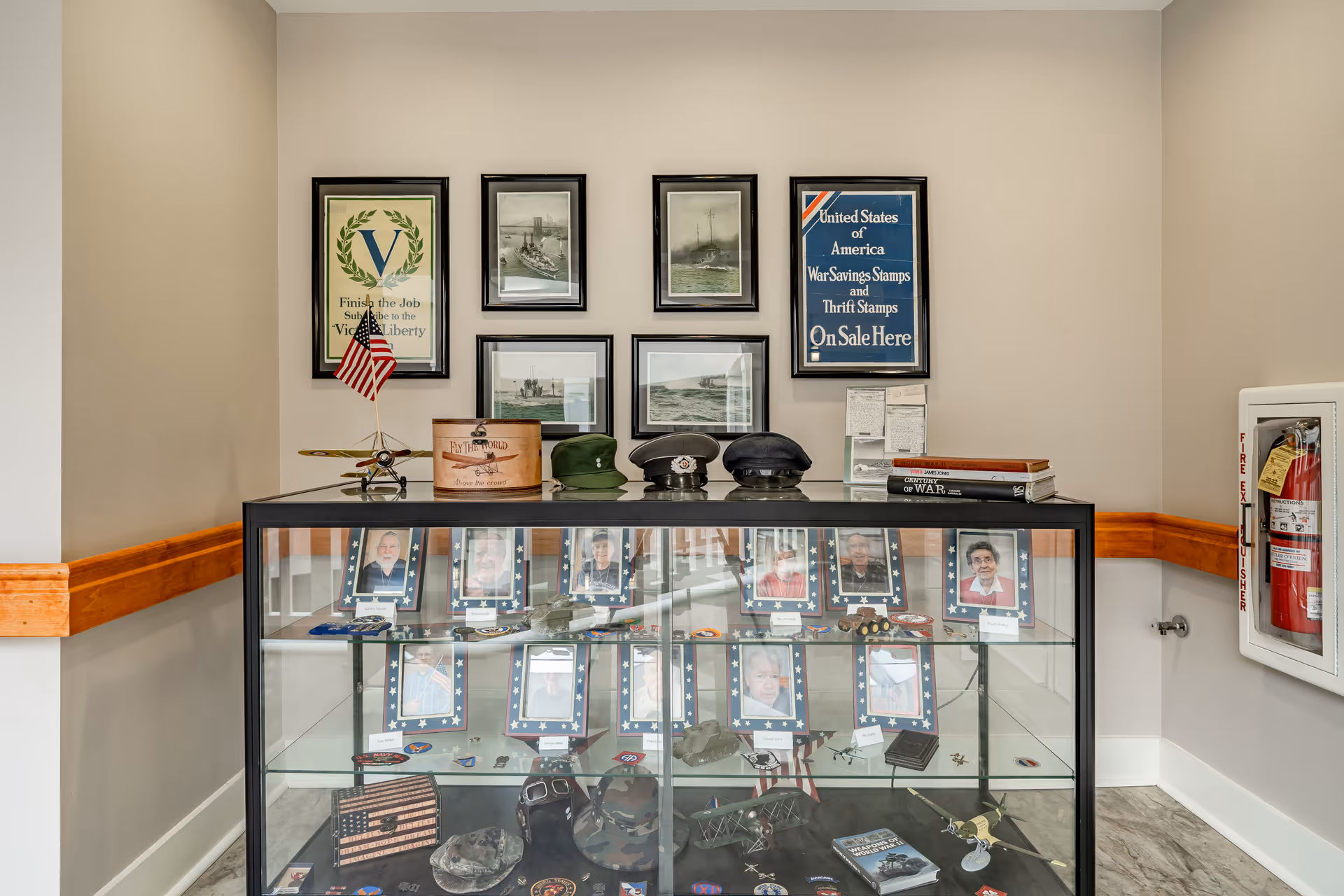 A glass display case in a hallway corner featuring military memorabilia, including framed photos of veterans, military hats, model airplanes, and books. Above the case, six framed vintage posters and photographs related to war and patriotism are hung on a beige wall. A fire extinguisher is mounted on the right wall.
