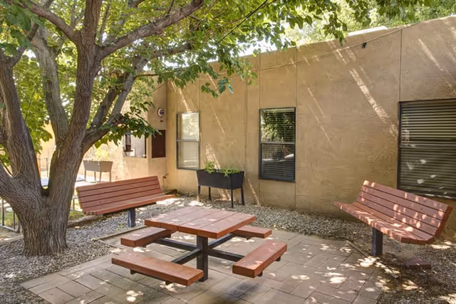 Outdoor seating area with a wooden picnic table and benches under a large tree, next to a beige building with windows and a small planter box.