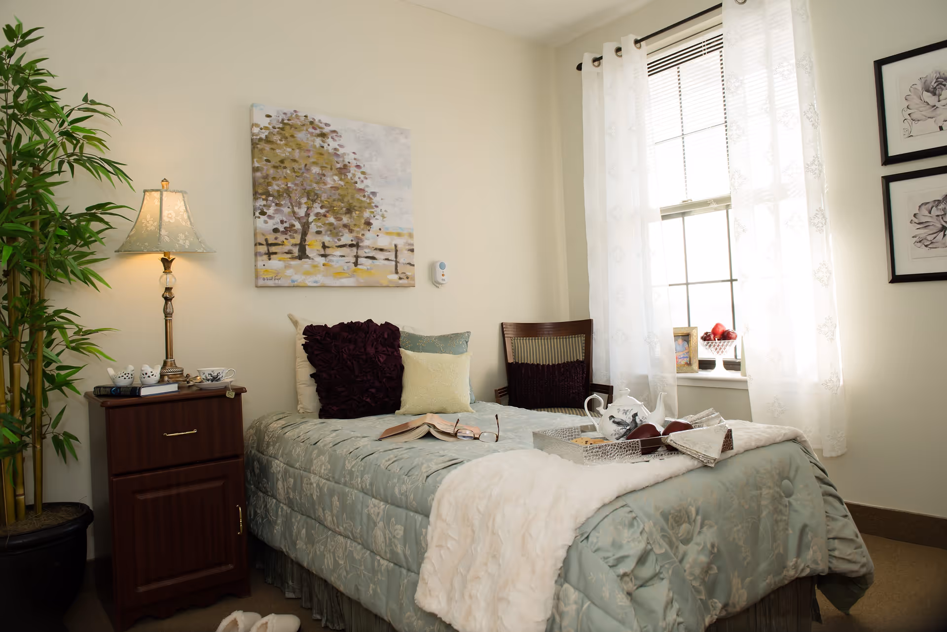 A cozy bedroom in a senior living facility with a single bed covered in a light blue floral comforter and multiple pillows. There is a wooden nightstand with a lamp, books, and decorative items. A large potted plant is next to the nightstand. A window with sheer white curtains allows natural light to fill the room. On the wall above the bed is a painting of trees and a fence. A chair with a cushion is placed near the window, and a tray with a teapot and apples rests on the bed.