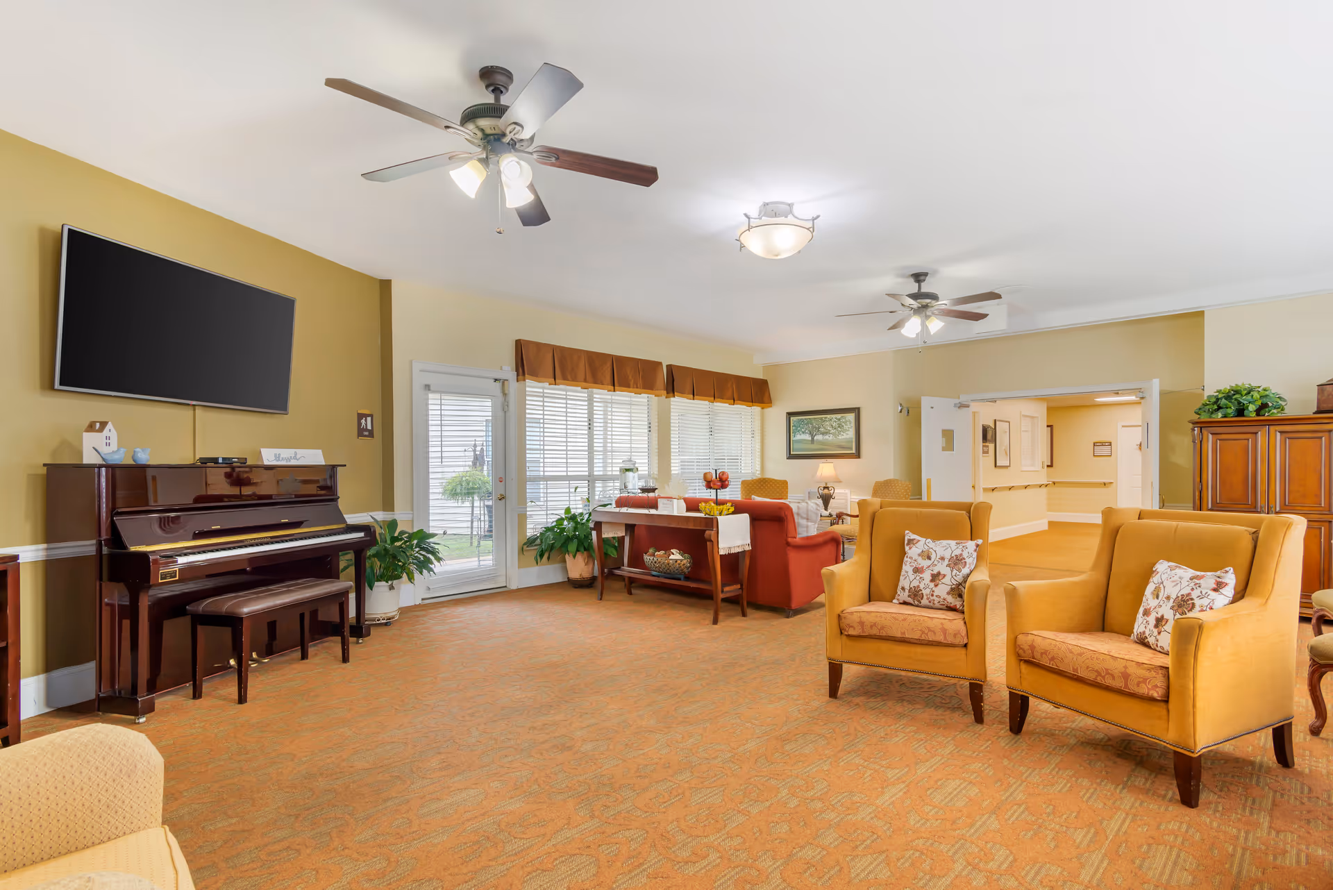 A spacious living room area with two yellow armchairs with floral cushions in the foreground, a red sofa and armchairs near a window with blinds and valance, a wooden piano with a bench beneath a mounted flat-screen TV, ceiling fans with lights, a wooden cabinet, and a door leading outside.