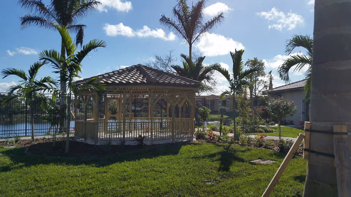 Wooden gazebo with a tiled roof on a grassy lawn surrounded by palm trees and a lake in the background.