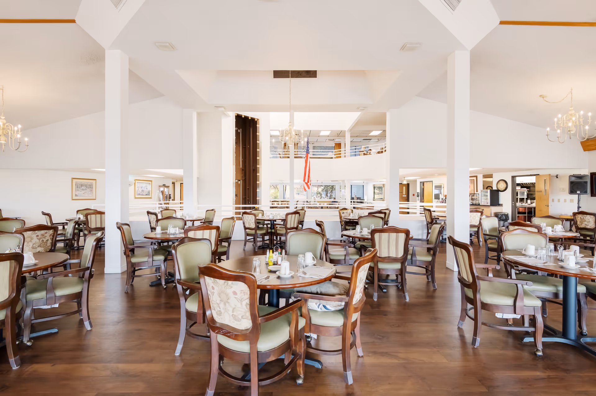 A spacious dining room with multiple round wooden tables and cushioned chairs arranged neatly. The room features white walls, wooden flooring, chandeliers, and an American flag hanging in the center background. There are windows and an open balcony area visible in the background.