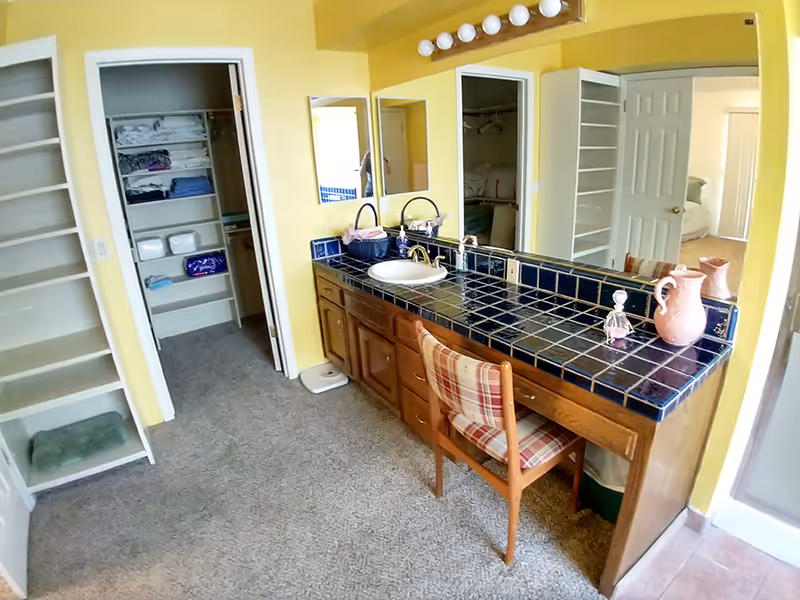 Carpeted dressing area with a blue-tiled vanity and sink, mirror lights, a plaid chair, and open closets against yellow walls.