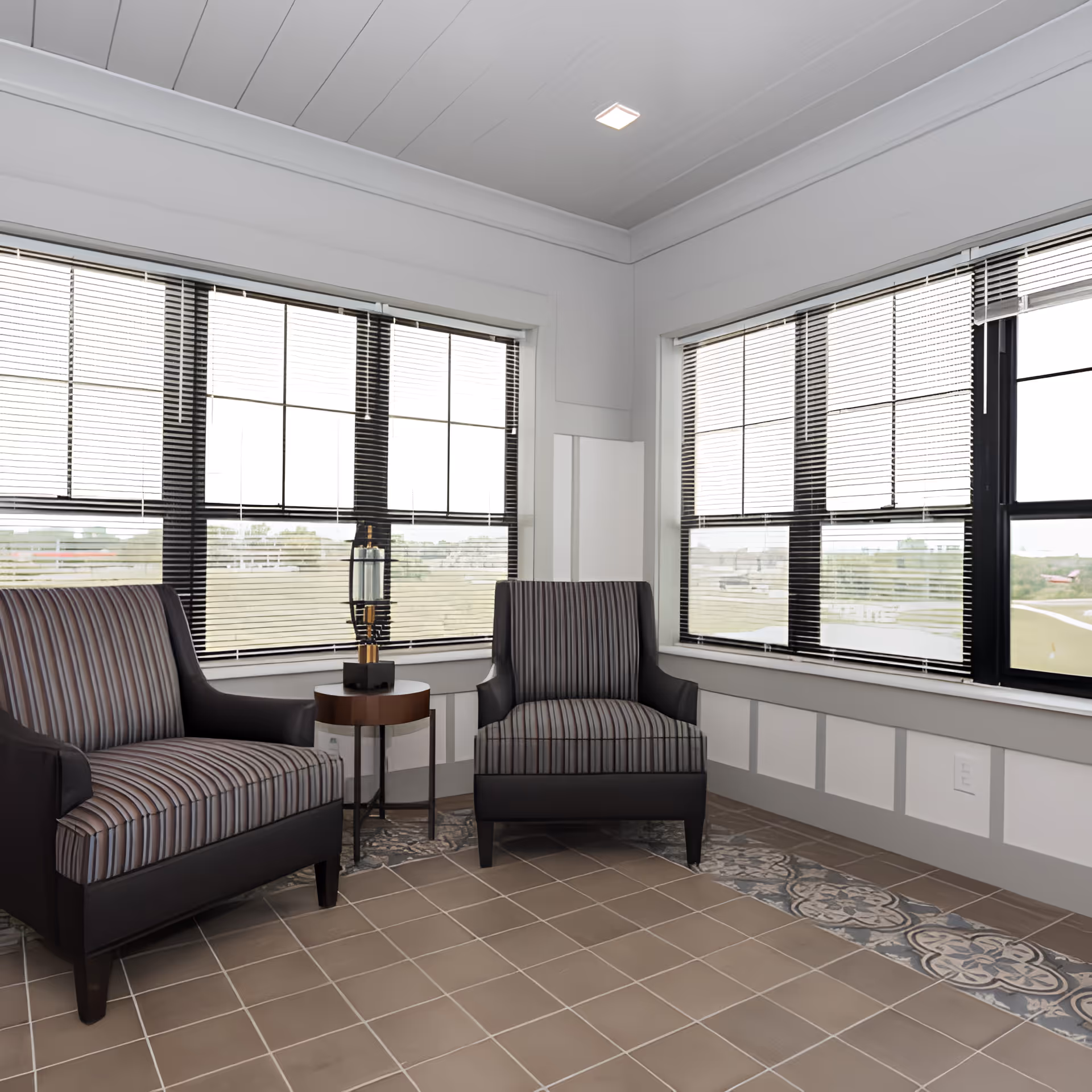 A bright corner seating area with two striped armchairs and a small round side table with a decorative lamp between them. The space has large windows with blinds, white paneled walls, and a tiled floor with a decorative border.