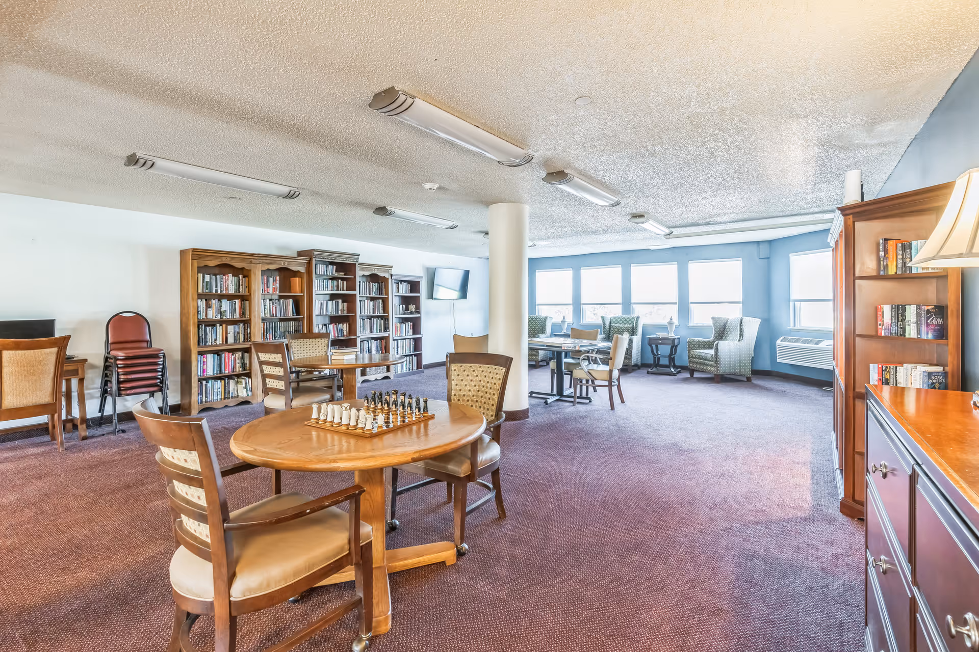 A spacious senior living common area with multiple wooden tables and chairs, a chessboard on one table, bookshelves filled with books along the wall, a flat-screen TV mounted in the corner, and large windows letting in natural light. The room has carpeted floors and a cozy, inviting atmosphere.