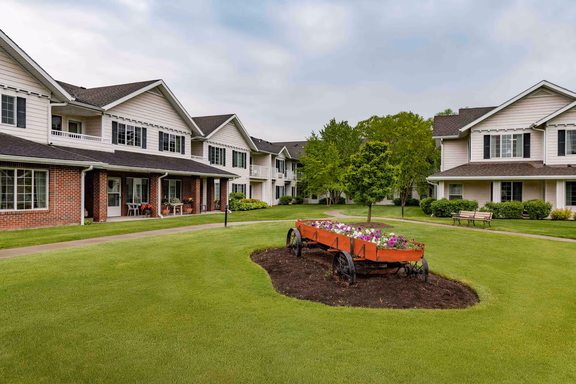 A well-maintained outdoor courtyard area of a senior living facility with green grass, a flower bed in a decorative wagon, trees, benches, and two-story residential buildings with balconies and porches.