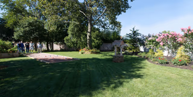 Sunlit landscaped lawn with a large tree, flower beds, a decorative wishing well, a bocce court, and a small group of people at the left edge.