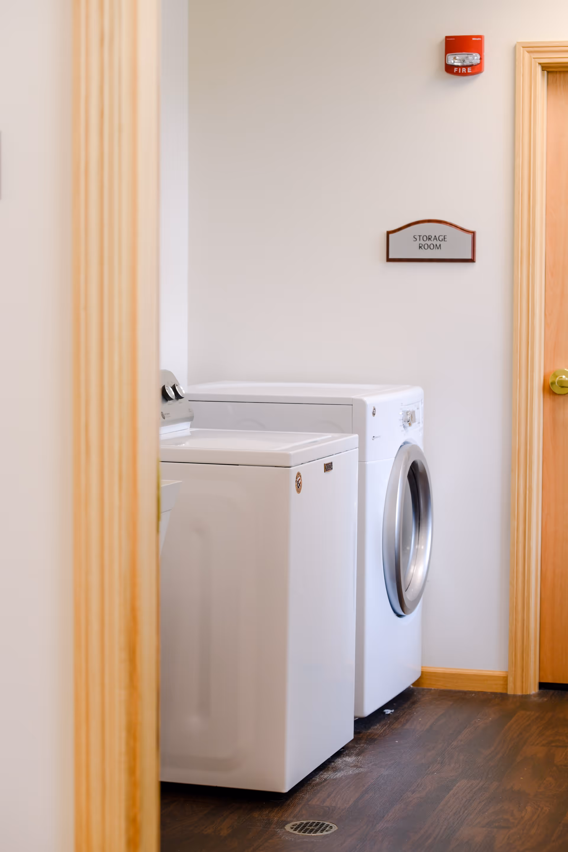 Laundry room with a white top-loading washing machine and a white front-loading dryer on a dark wooden floor. A sign on the wall reads 'Storage Room' and a red fire alarm is mounted above it.