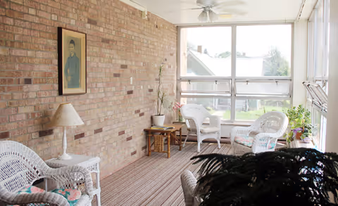 A bright sunroom with large windows letting in natural light. The room features white wicker chairs with floral cushions, a wicker side table with a lamp, a small wooden table with a potted plant, and a framed portrait hanging on a brick wall. There are additional plants near the windows and a ceiling fan overhead.