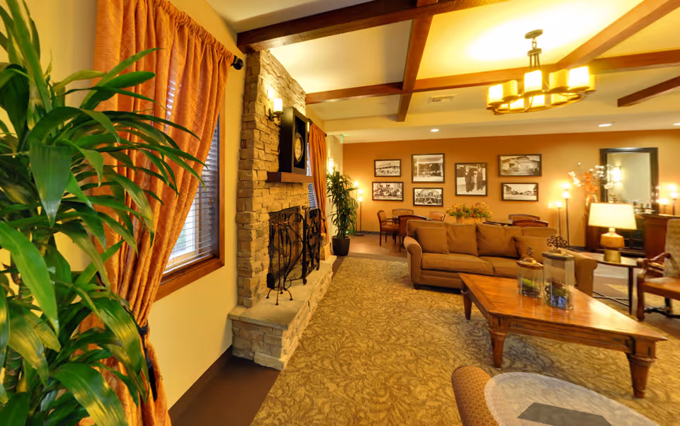 A cozy senior living facility common area with a stone fireplace, a large clock above it, and orange curtains on the windows. The room features a brown sofa, wooden coffee table with decorative items, framed black and white photos on the wall, several chairs, and warm lighting from lamps and ceiling fixtures. There are also green plants adding a touch of nature to the space.