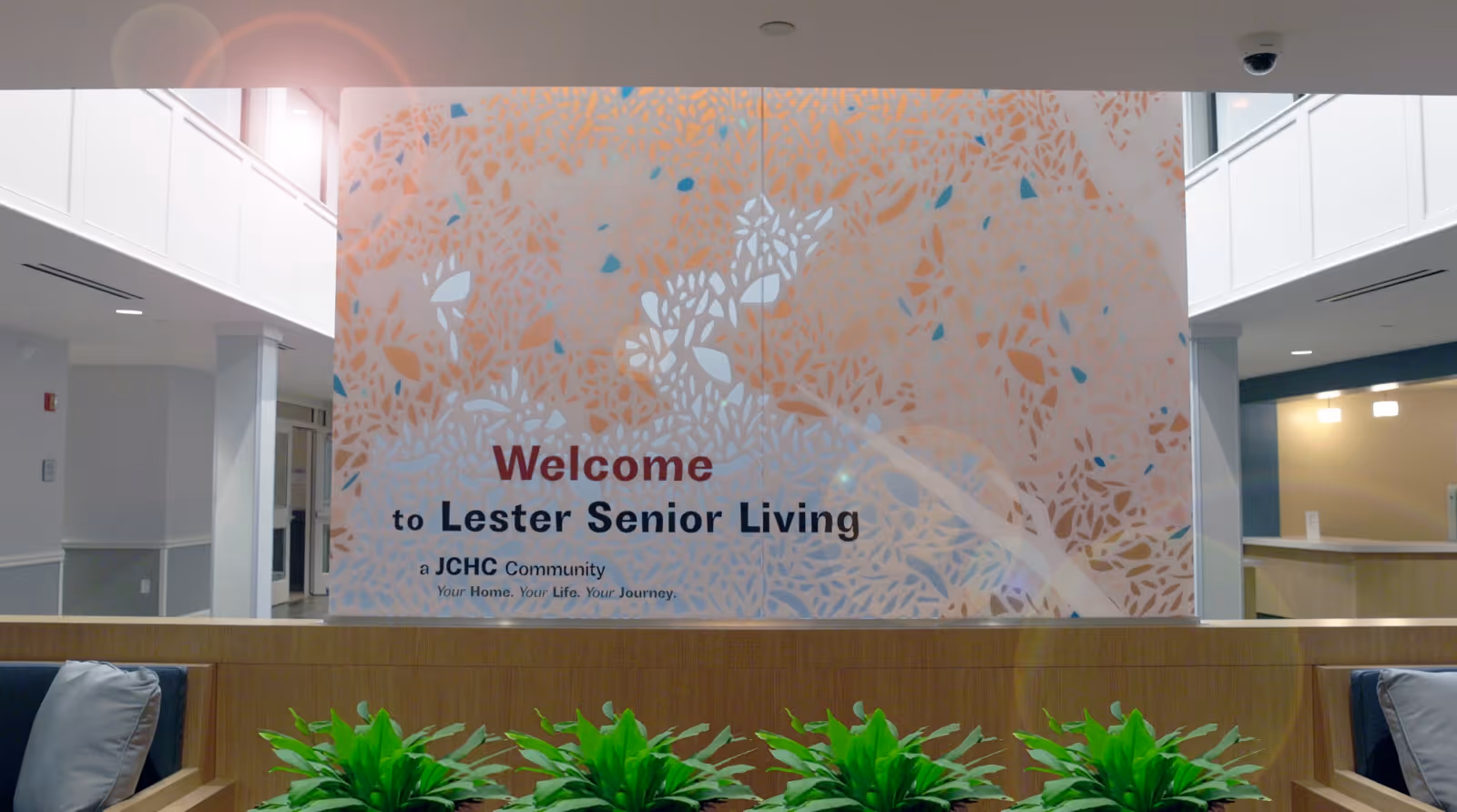 Interior lobby showing a decorative wall sign reading 'Welcome to Lester Senior Living' above seating and plants.
