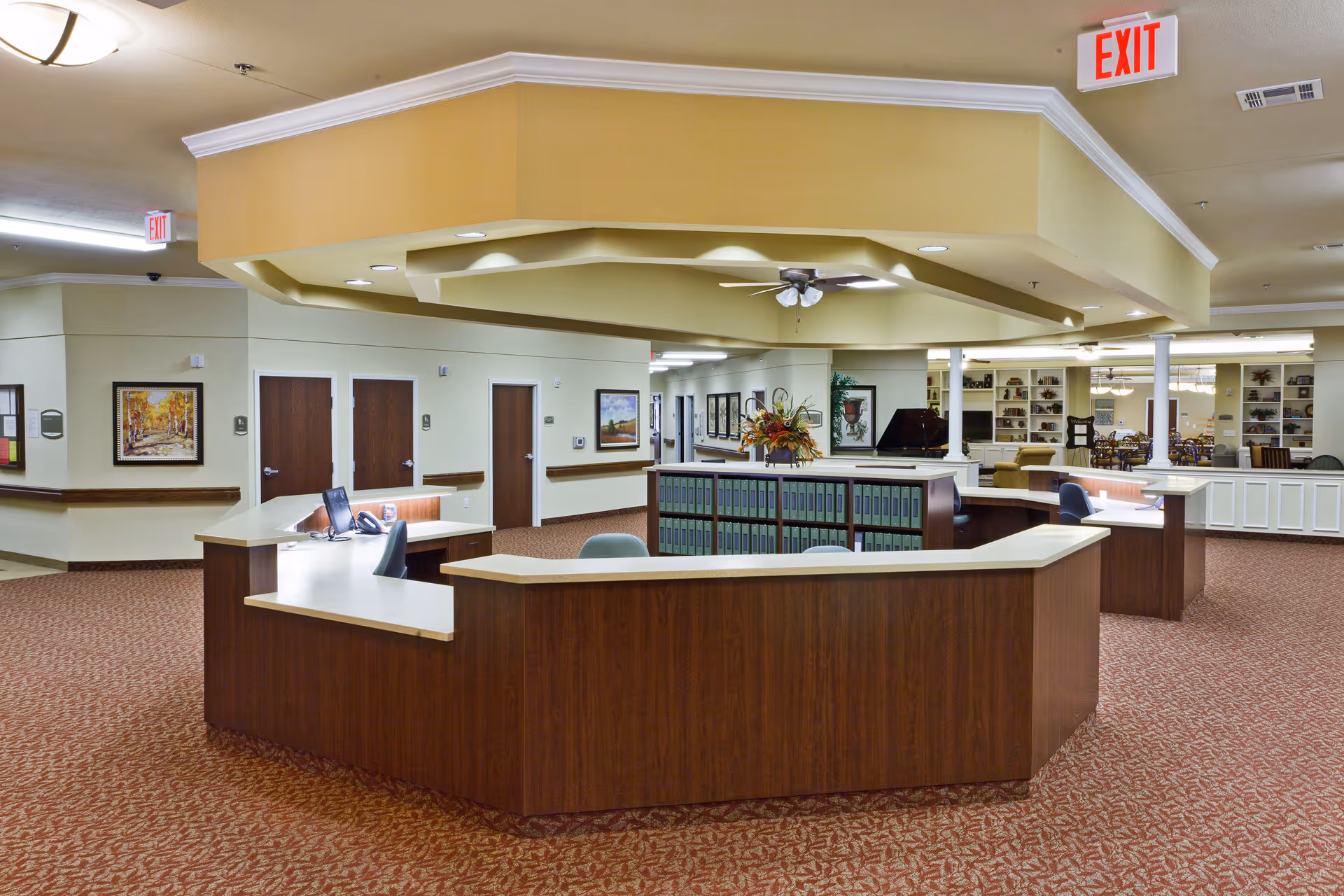 A spacious senior living facility lobby with a central wood reception desk, seating, bookshelves, and overhead lighting.