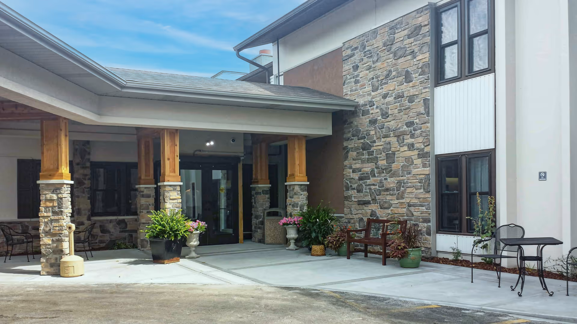 Entrance area of a senior living facility with stone and wood pillars, potted plants, a bench, and a small table with chairs under a covered walkway.