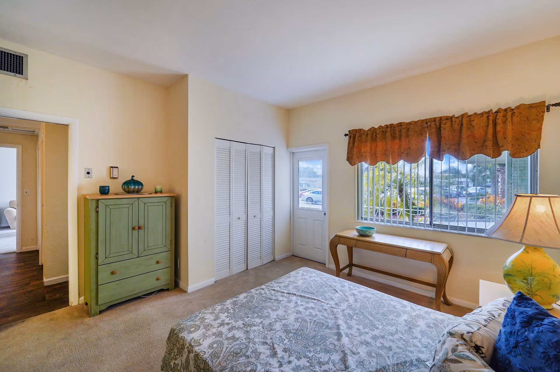 A bedroom with a bed covered in a patterned bedspread, a green cabinet with decorative items on top, a wooden table with a bowl near a window with orange curtains, and a lamp on a bedside table. The room has beige walls and carpeted floor, with a door and closet with white louvered doors.