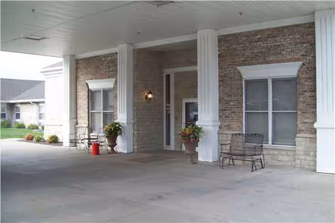 Covered entrance with brick building, tall columns, and seating near potted plants.