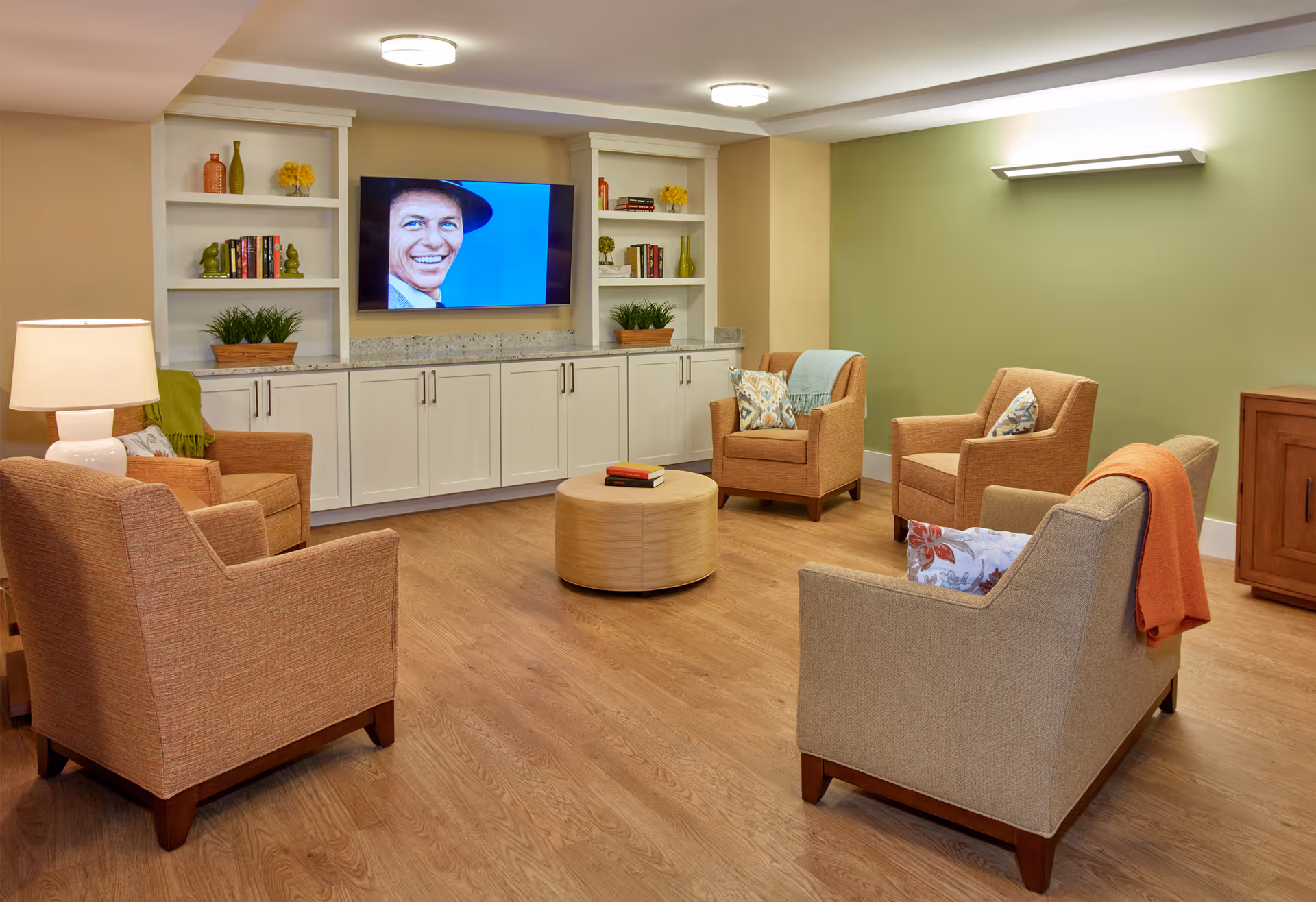 A cozy senior living common area with four upholstered armchairs arranged around a round wooden coffee table. The room features wood flooring, a green accent wall, built-in white cabinets with shelves holding decorative items and books, and a flat-screen TV mounted on the wall displaying a smiling man wearing a hat. There is a table lamp on the left and a wooden cabinet on the right.