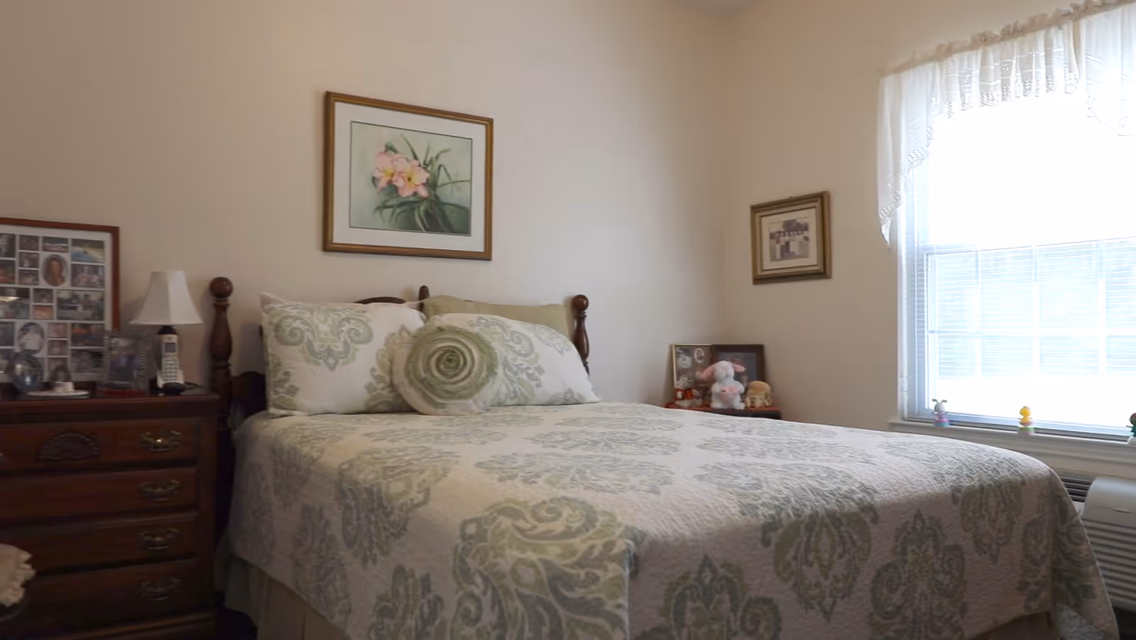 A neatly made bedroom with a patterned bed, wooden nightstand, framed pictures, and a sunlit window.
