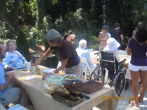 A group of people gathered outdoors around a table with food, including a woman serving food and others sitting or standing nearby, some in wheelchairs, surrounded by trees and greenery.