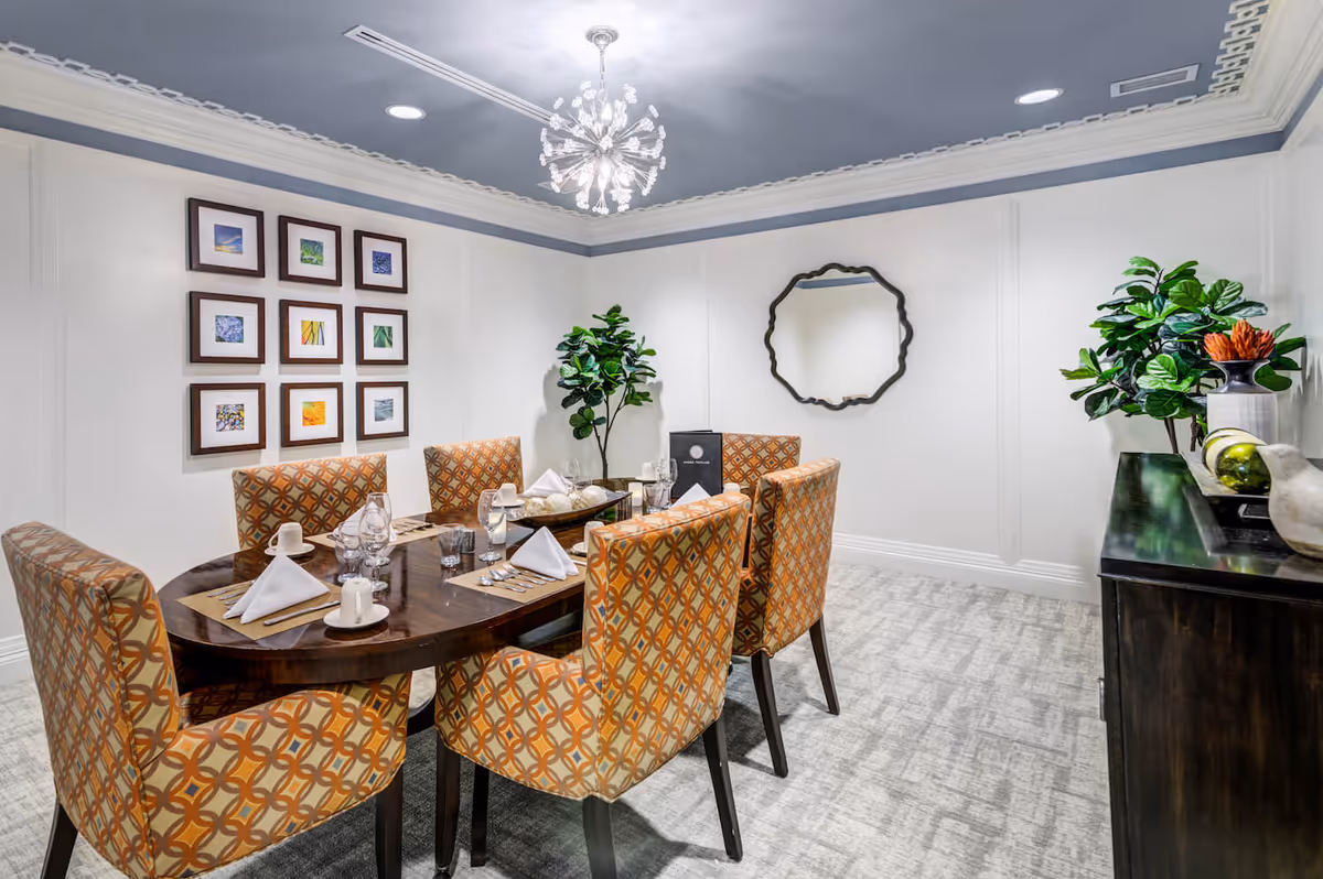 A well-lit dining room with a dark wooden oval table surrounded by six patterned orange and beige chairs. The table is set with white napkins, cups, glasses, and silverware. The walls are white with decorative molding, featuring a set of nine framed pictures on one side and a decorative mirror on another. There are two large green potted plants and a dark wooden sideboard with decorative items. The ceiling is painted blue with a modern chandelier hanging in the center.