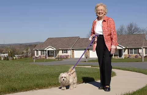 An elderly woman wearing a red patterned jacket, white shirt, and black pants is walking a small white dog on a purple leash along a sidewalk in a residential area with single-story houses and a clear blue sky.