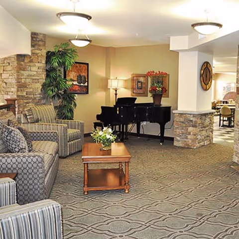 A cozy living room area with patterned carpet, two striped armchairs, a patterned sofa, a wooden coffee table with a flower arrangement, a black grand piano with a flower pot on top, a floor lamp, framed artwork on the walls, and a large potted plant near a stone accent wall.