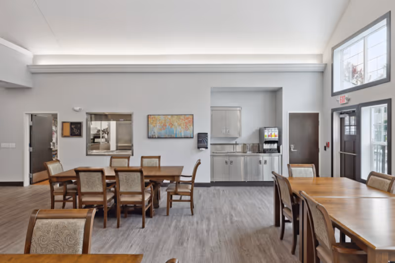 Dining room with wooden tables and chairs, a serving counter and drink dispenser against a light wall and large windows.