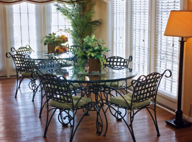 A bright dining area with two round glass tables surrounded by black metal chairs with green cushions. The room has large windows with white blinds allowing natural light to fill the space. There are green plants on the tables and a tall floor lamp in the corner.