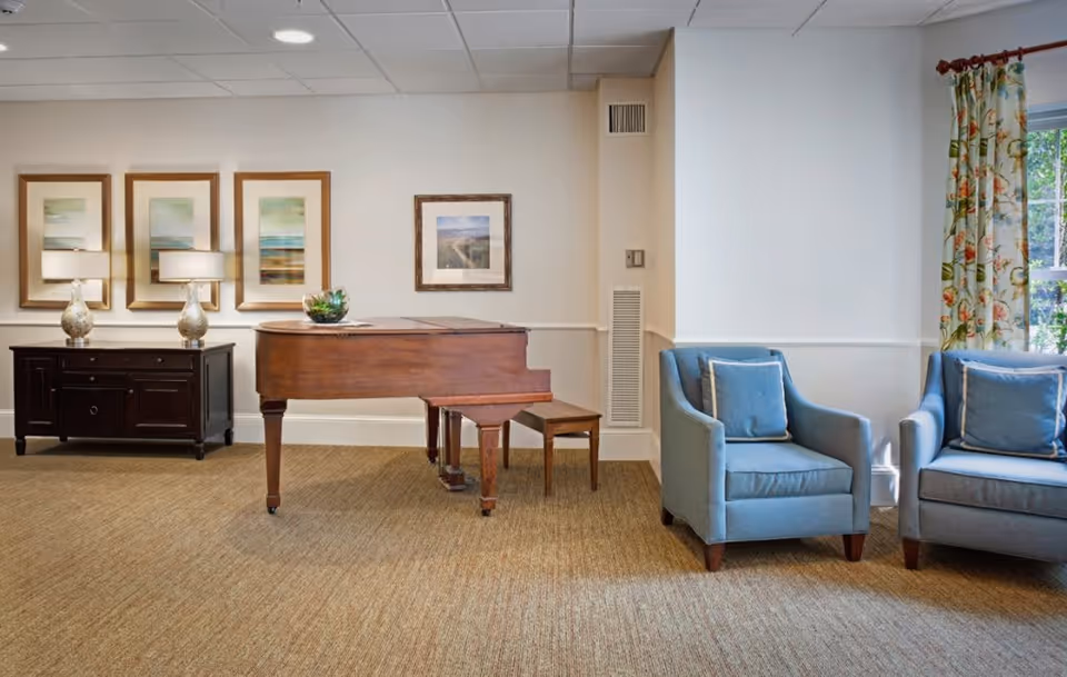 A cozy interior room featuring a wooden grand piano with a matching bench, two blue upholstered armchairs with cushions, a dark wooden sideboard with two decorative lamps, and framed artwork on the walls. A window with floral curtains allows natural light to enter the room.