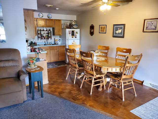Open-plan living and dining area featuring a wooden dining table with chairs, a small kitchen in the background, and a recliner.